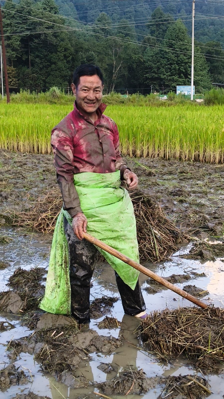 A smiling volunteer in muddy clothes holding a hoe in a paddy field